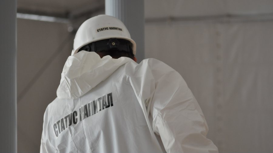 Сотрудник со спины в каске и комбинезоне с надписью «СТАТУС КАПИТАЛ» в павильоне