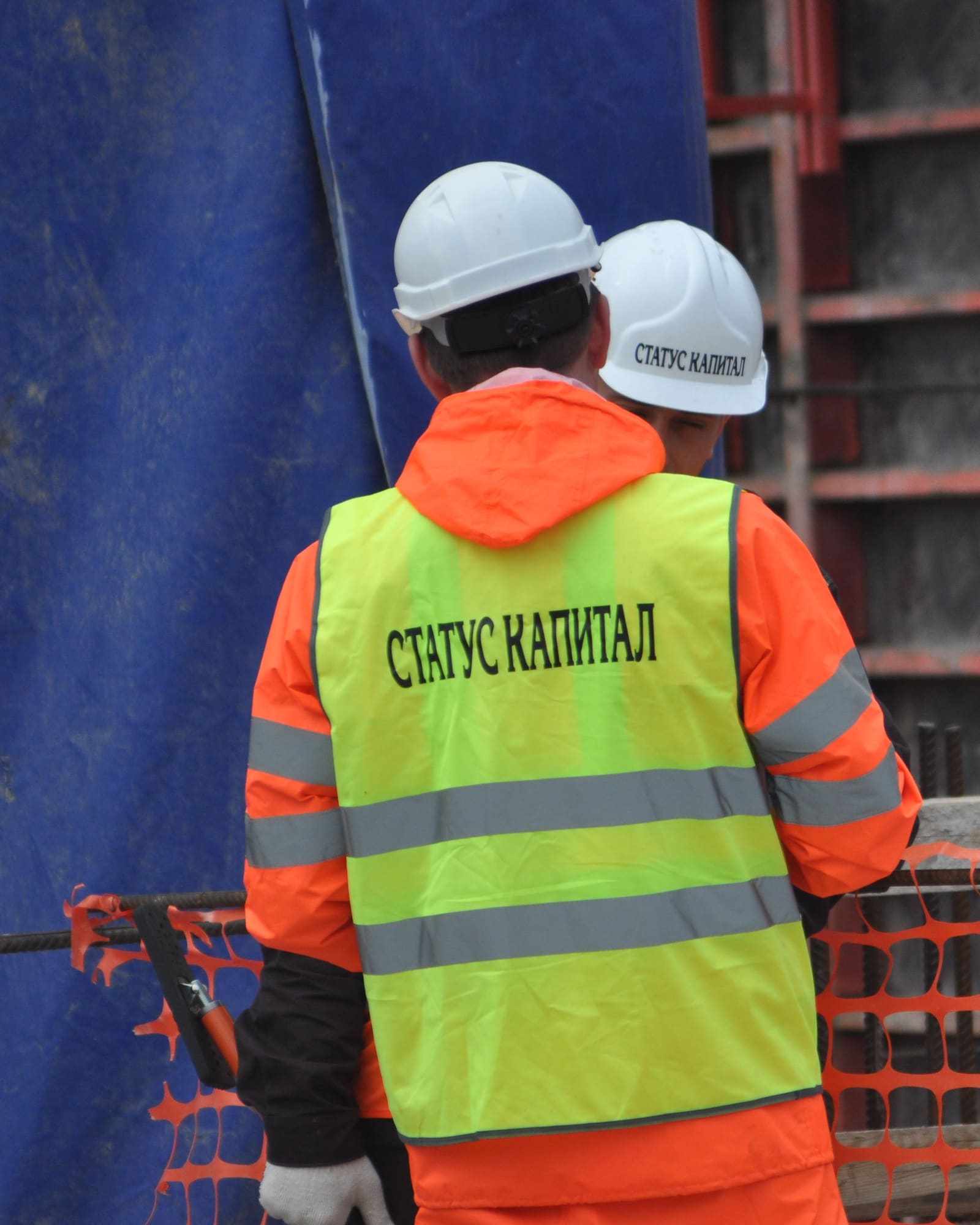 Два наших инженера со спины, надпись «Статус Капитал» на жилете и каске, перед ними сетчатое инвентарное ограждение рабочей зоны, за ним щиты опалубки
