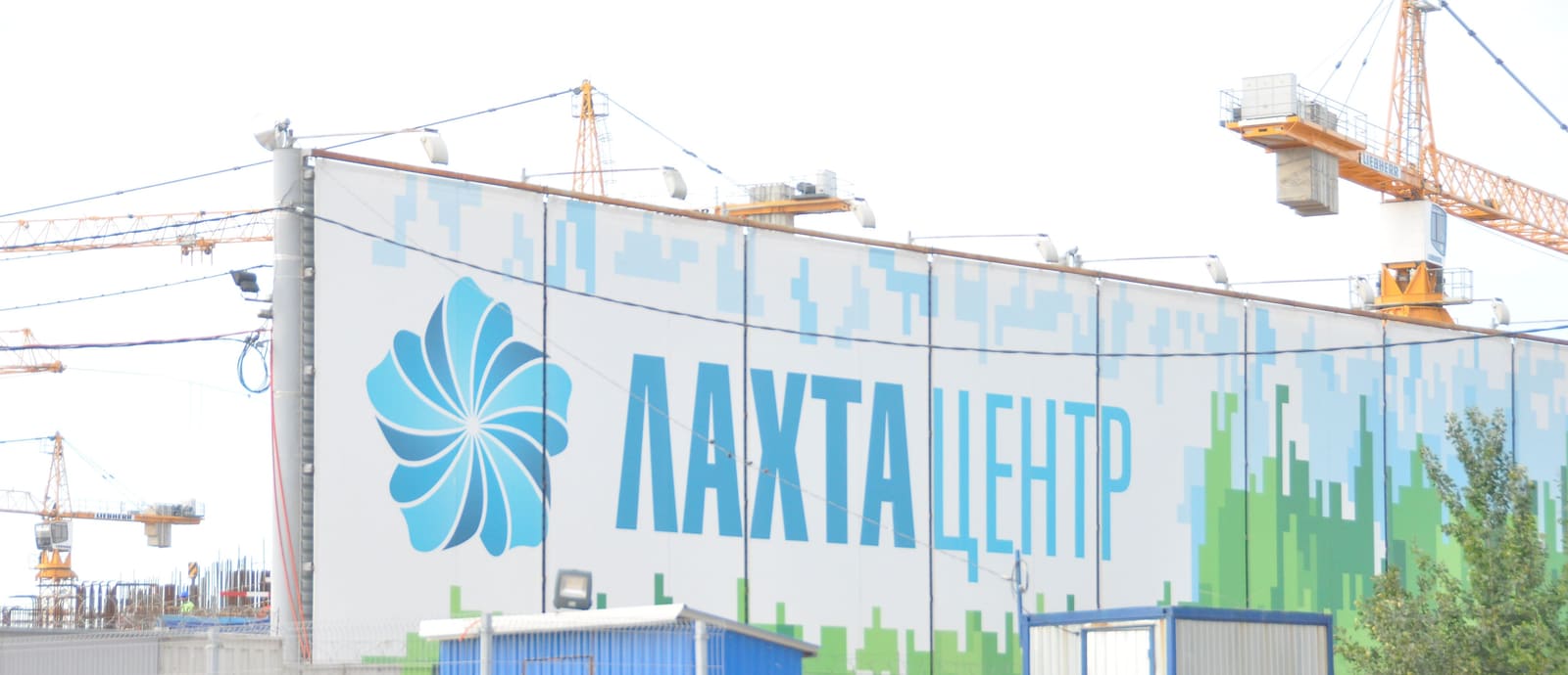 Забор стройплощадки с надписью «Лахта Центр», за ним башенные краны